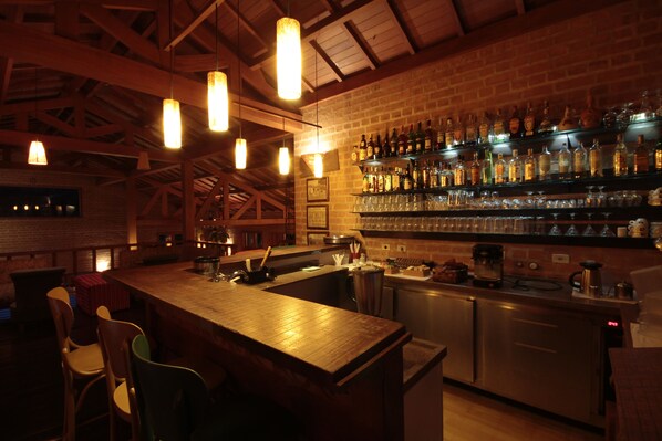 Una zona de bar con vigas de madera, luces colgantes y estantes llenos de botellas y copas.