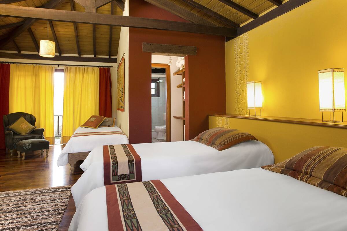 A hotel room with two beds, a wooden ceiling, a yellow wall, and a window with yellow curtains.