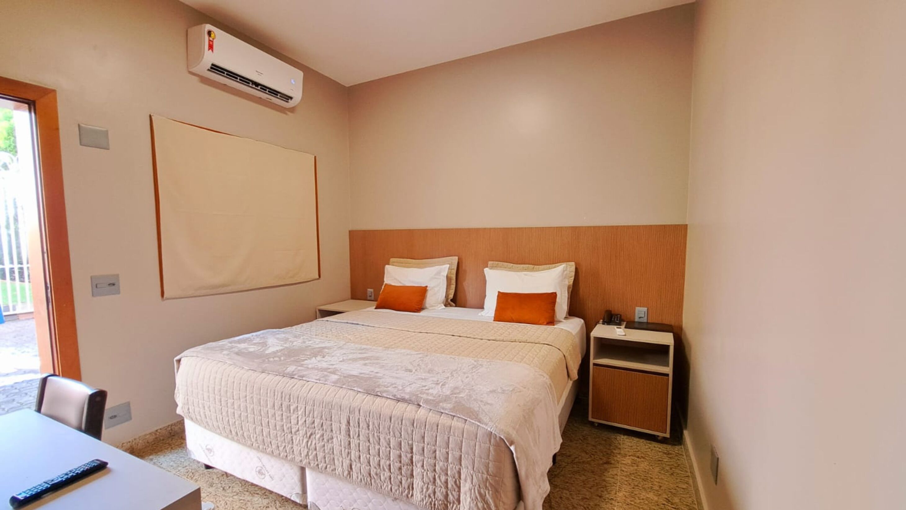 A hotel room with a bed, bedside tables, a desk, and an air conditioning unit.