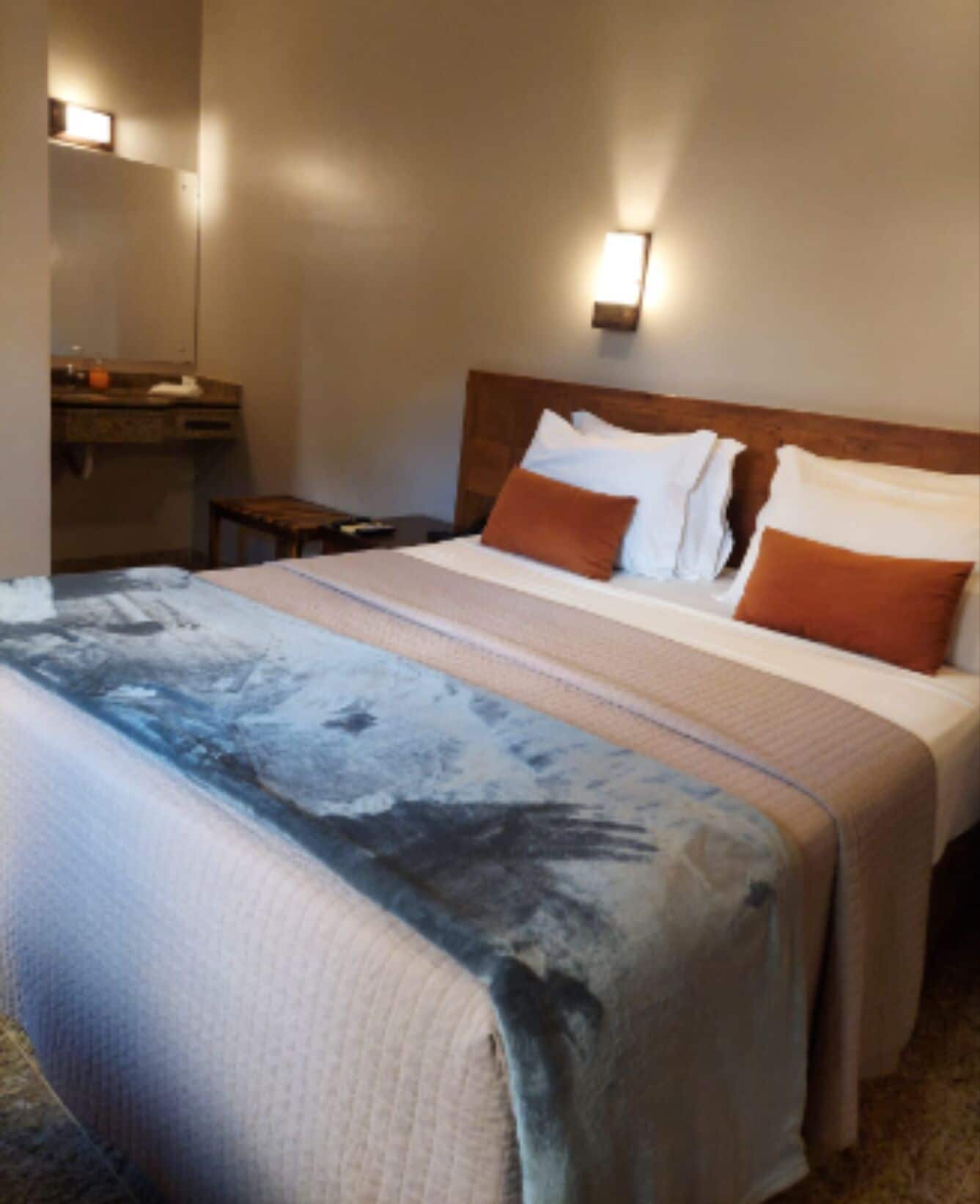 A hotel room with a bed, a nightstand, a mirror, and a wall-mounted light fixture.