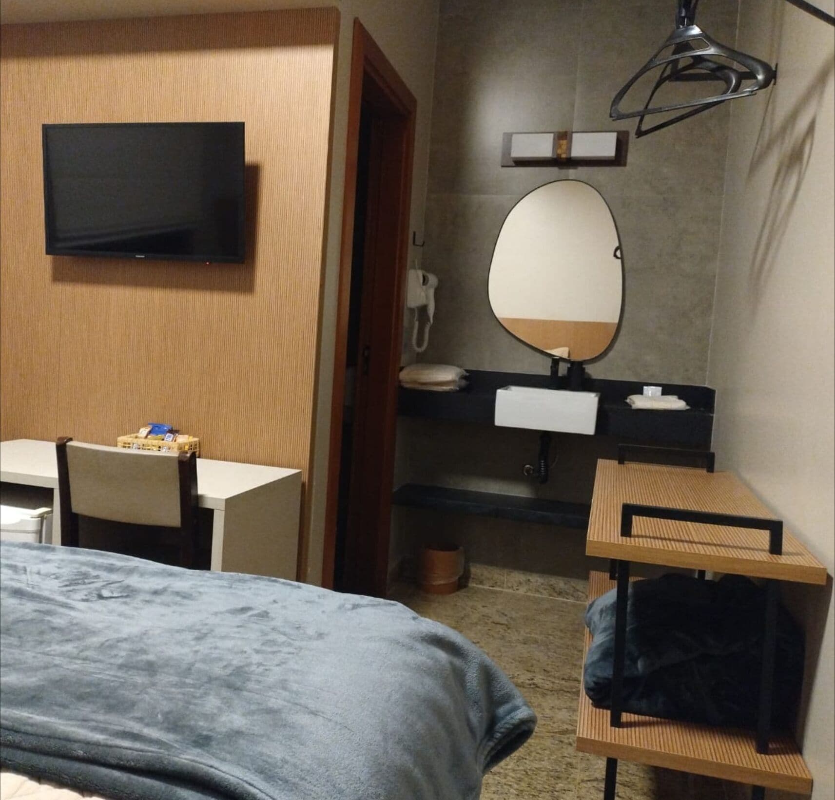 Habitación de hotel con cama, escritorio, televisor, espejo, lavamanos y perchero.