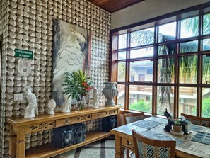 Una stanza con parete strutturata, un'ampia finestra vista palme, un tavolo in legno con tovaglioli decorati e uno scaffale ornamentale con diversi oggetti.
