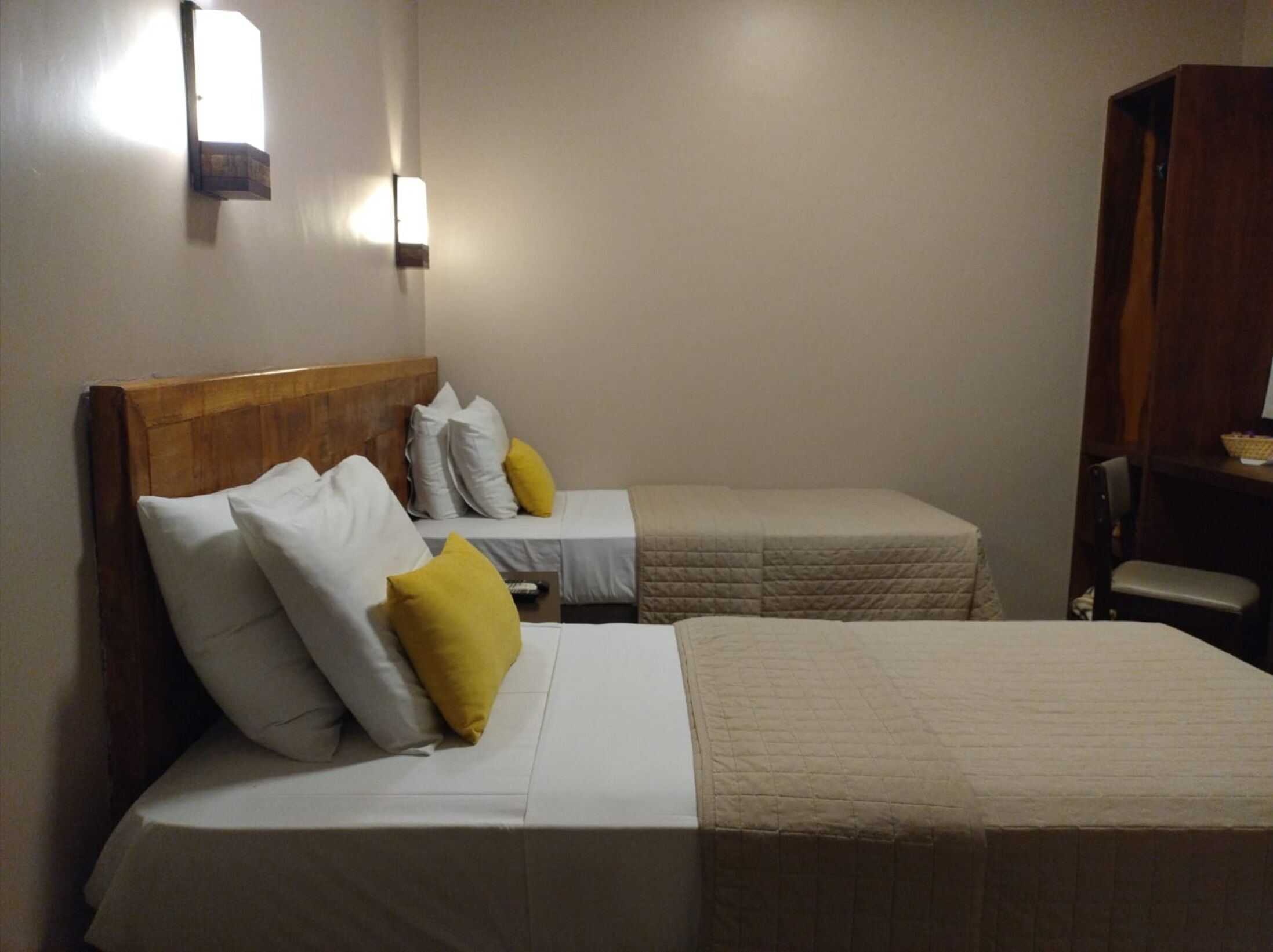 Habitación de hotel con dos camas, cada una con ropa blanca y cabeceros de madera. Se observan dos lámparas fijadas a la pared y un armario de madera.