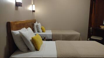 Habitación de hotel con dos camas, cada una con ropa blanca y cabeceros de madera. Se observan dos lámparas fijadas a la pared y un armario de madera.