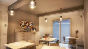 Ein moderner Essbereich mit weißen Tischen und Stühlen, Pendelleuchten, einem farbenfrohen Gemälde mit Früchten an der Wand und einem Fenster mit Jalousien.