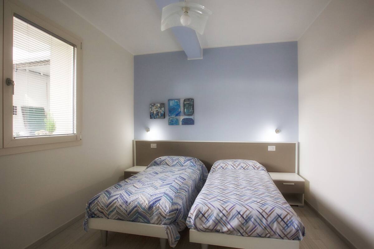 Un dormitorio con dos camas individuales, una ventana con persianas, un ventilador de techo e iluminación empotrada en la pared.