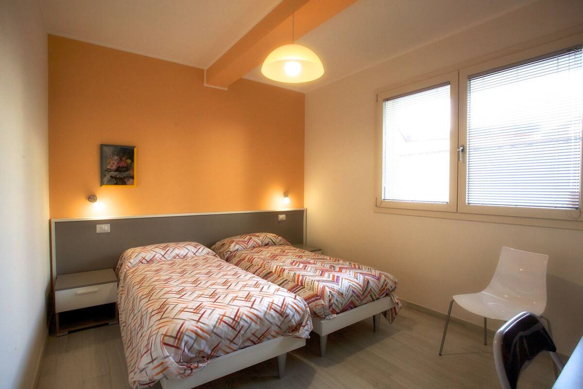 Un dormitorio con dos camas, una silla, una ventana con persianas y un cuadro en la pared.