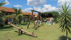 Une aire de jeux avec des balançoires en bois et une balançoire à bascule, située dans un espace verdoyant agrémenté de palmiers et d’un bâtiment dont le toit est recouvert de tuiles rouges.