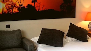 A bedroom with a bed, two pillows, a grey armchair, and a large wall painting of a sunset with silhouettes of trees. - Résidence Marina Beach (Nouméa)