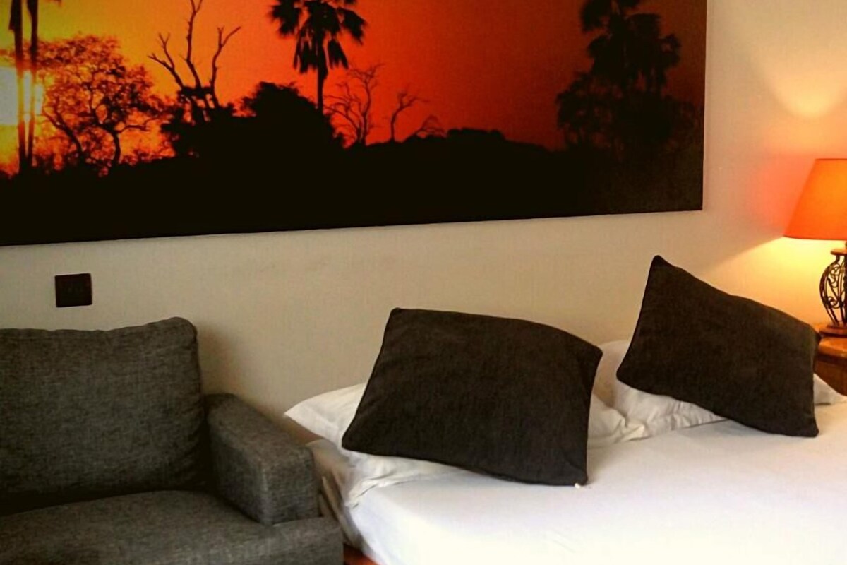 A bedroom with a bed, two pillows, a grey armchair, and a large wall painting of a sunset with silhouettes of trees.