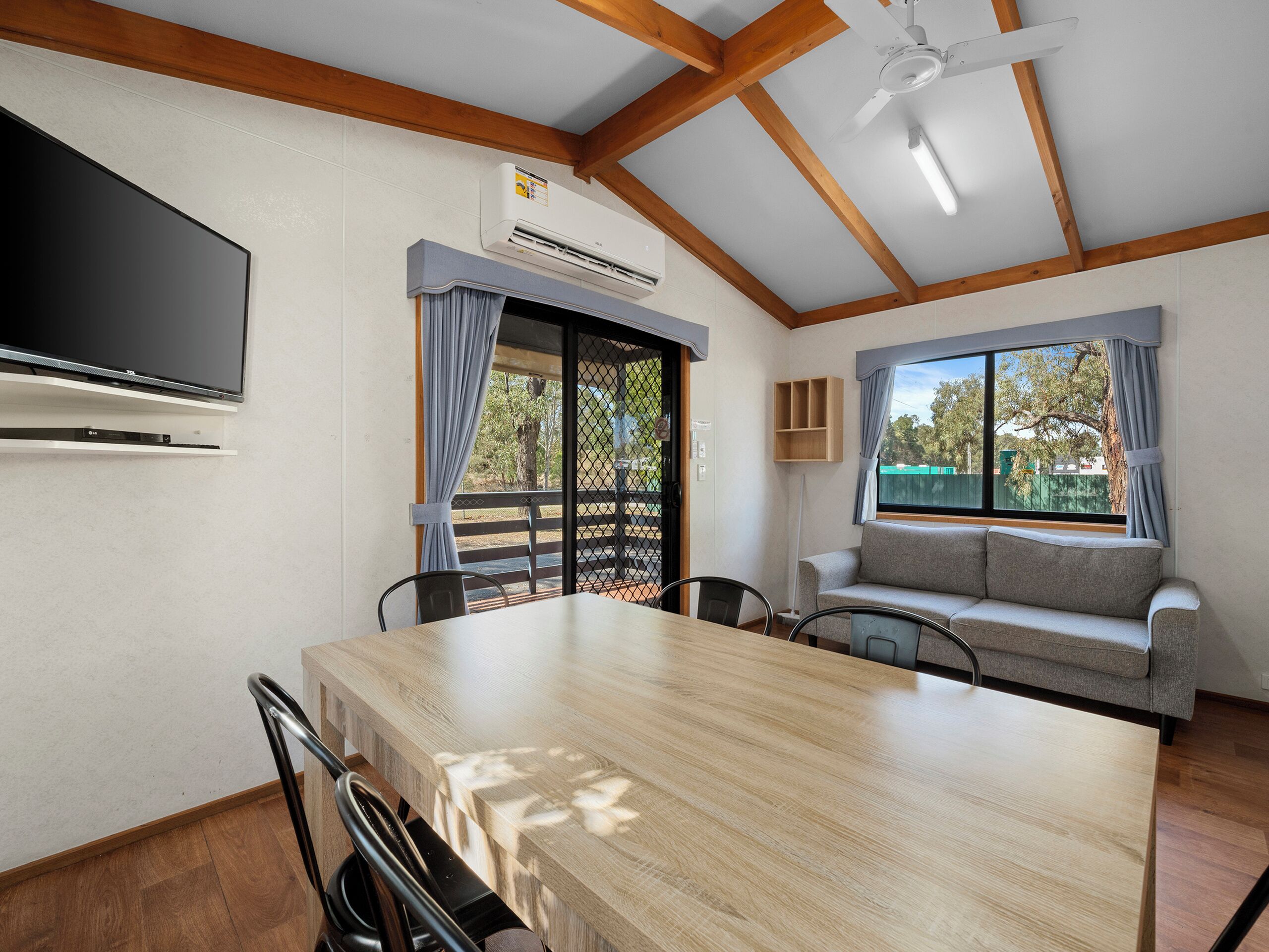 Una habitación con una mesa de comedor de madera, sillas, un sofá, un televisor montado en la pared y un equipo de aire acondicionado.