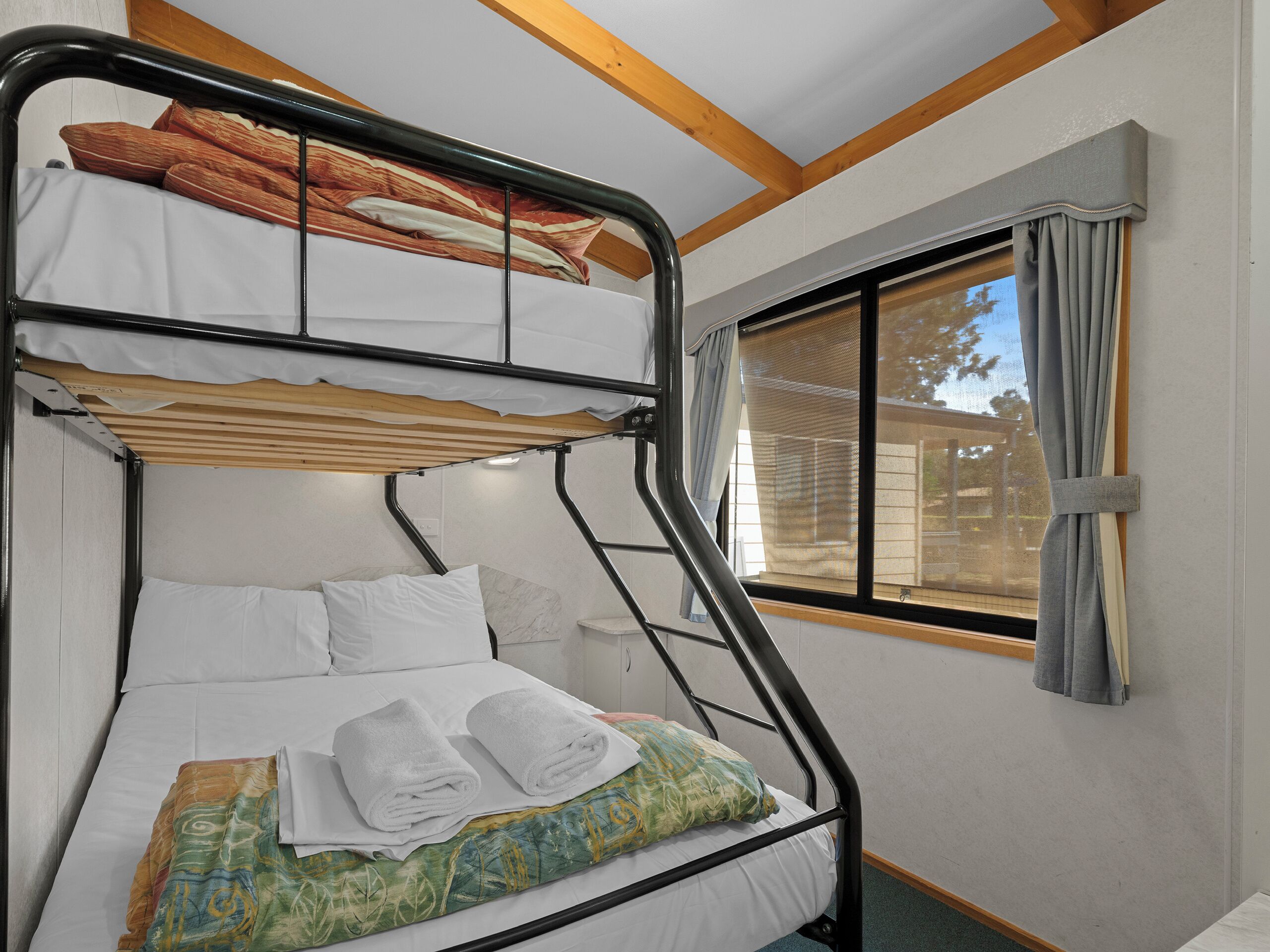 A bunk bed with white bedding and a colorful blanket, a window with curtains, and a wooden ceiling beam.