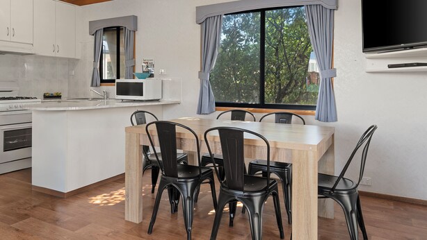 A modern kitchen with white cabinets, a microwave, and a stove. There is a dining area with a wooden table and black chairs. A large window with curtains is visible, and a flat-screen TV is mounted on the wall. - Wodonga Holiday Park (West Wodonga)