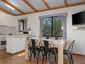 A modern kitchen with white cabinets, a microwave, and a stove. There is a dining area with a wooden table and black chairs. A large window with curtains is visible, and a flat-screen TV is mounted on the wall. - Wodonga Holiday Park (West Wodonga)