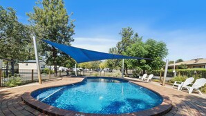 A swimming pool with a blue shade sail, surrounded by brick tiles and white lounge chairs. - Wodonga Holiday Park (West Wodonga)