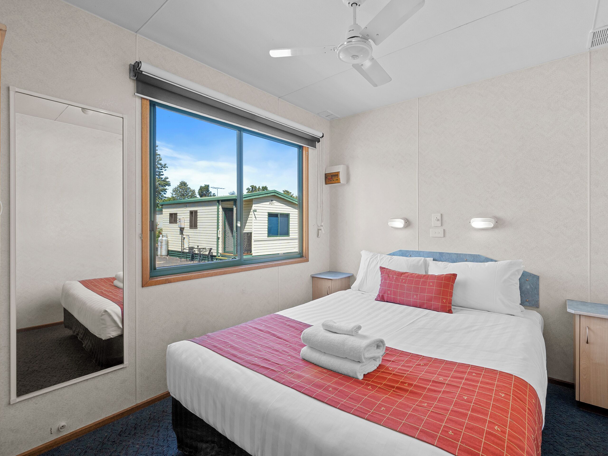 A neatly made bed with a red and white bedspread, a window with a view of a building outside, and a ceiling fan.