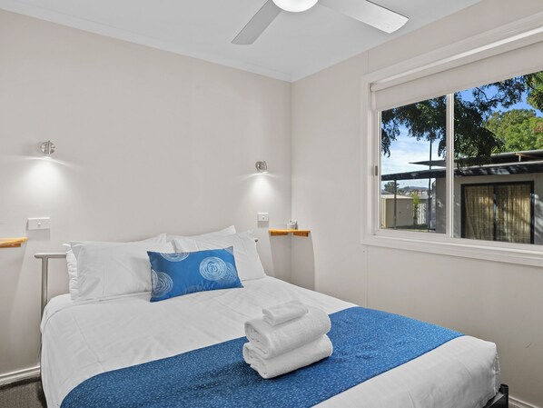 A neatly made bed with white and blue bedding, a window with a view of greenery, and a ceiling fan. - Wodonga Holiday Park (West Wodonga)