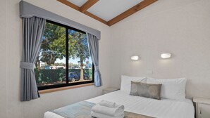 A neatly made bed with a patterned bedspread, a window with curtains, and a nightstand with a lamp. - Wodonga Holiday Park (West Wodonga)