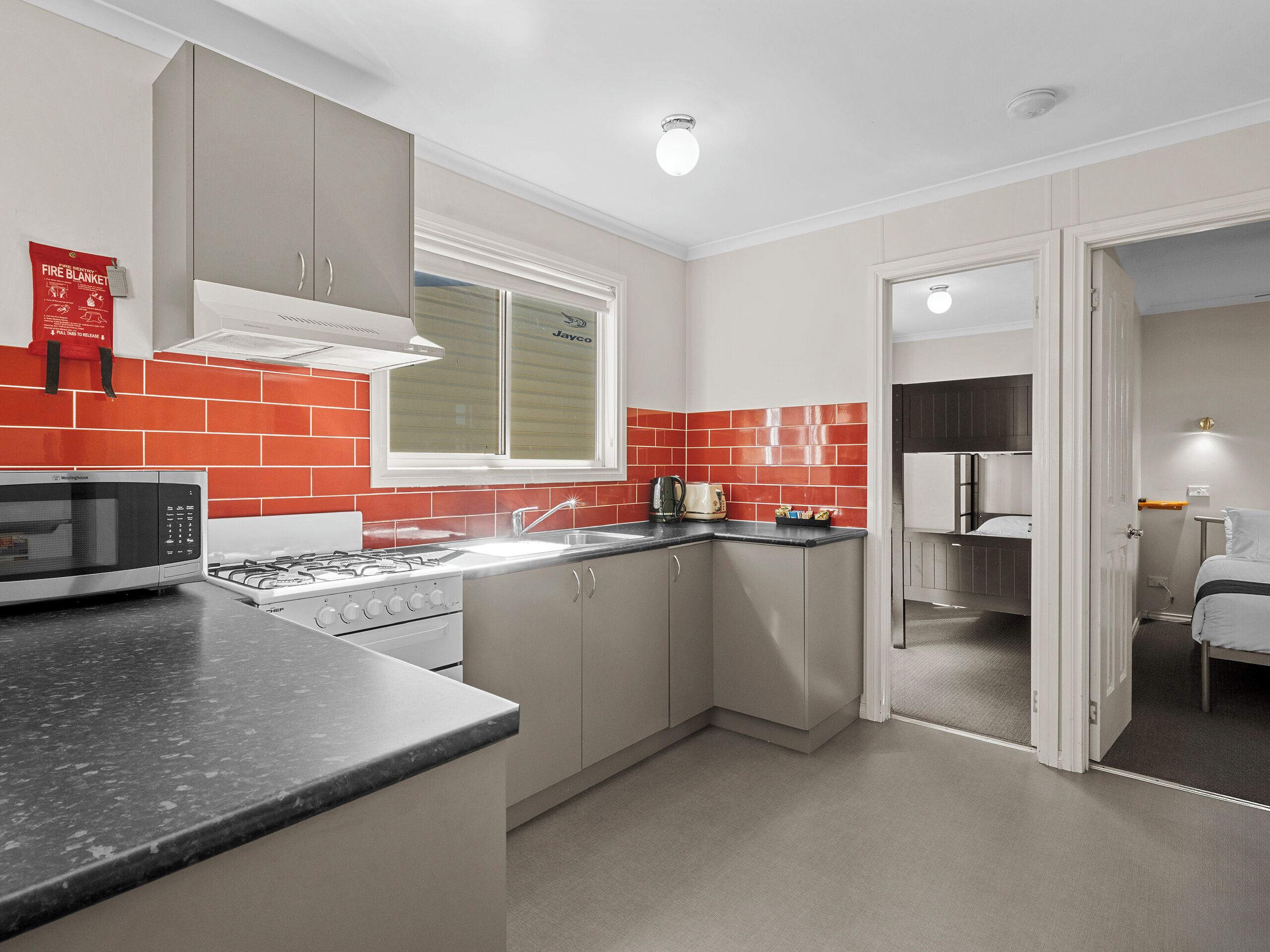 A compact kitchen with a microwave, stove, sink, and red-tiled backsplash.
