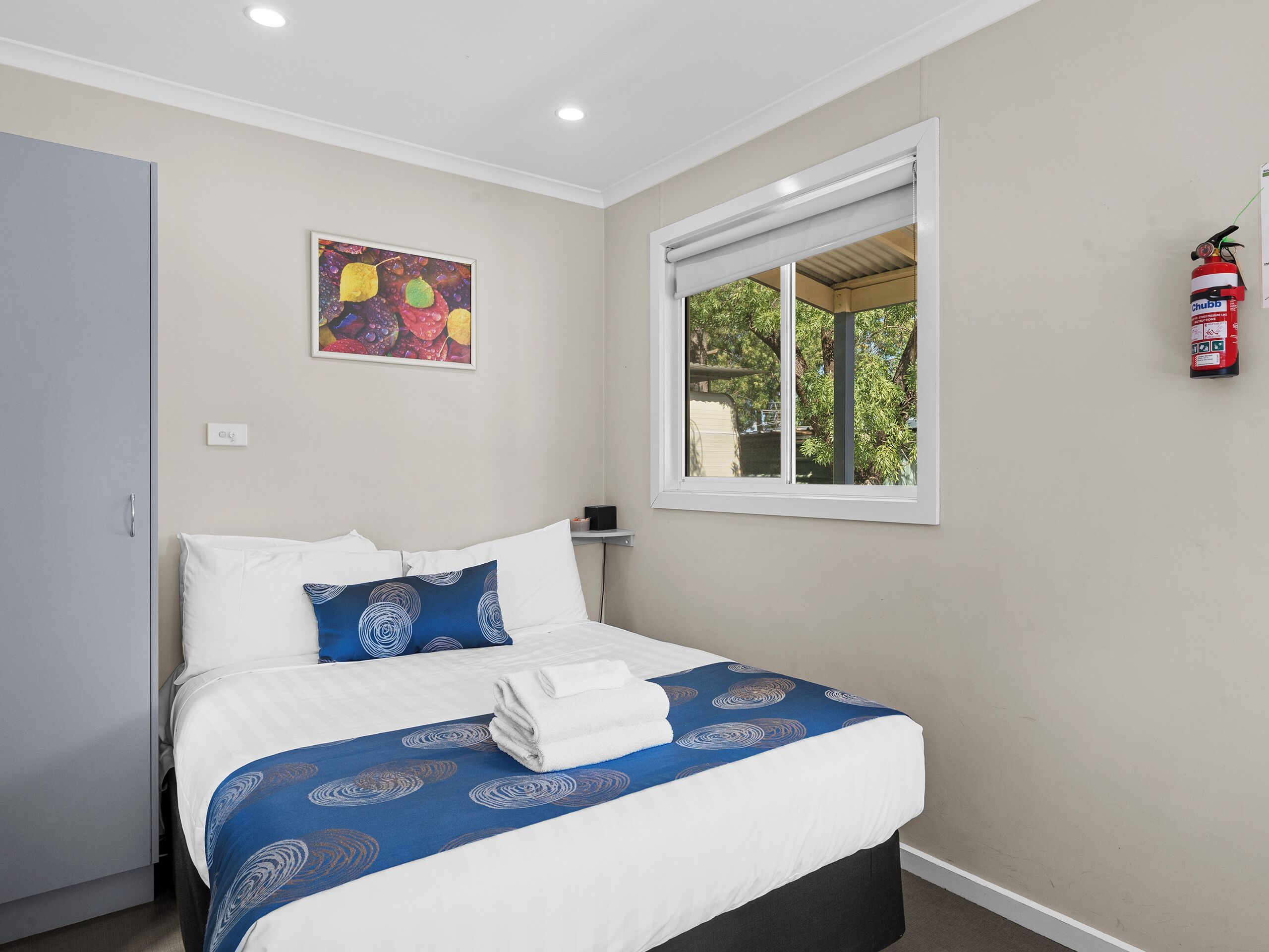 Una cama bien hecha con ropa de cama blanca y azul, un cuadro en la pared y una ventana con vistas al follaje.