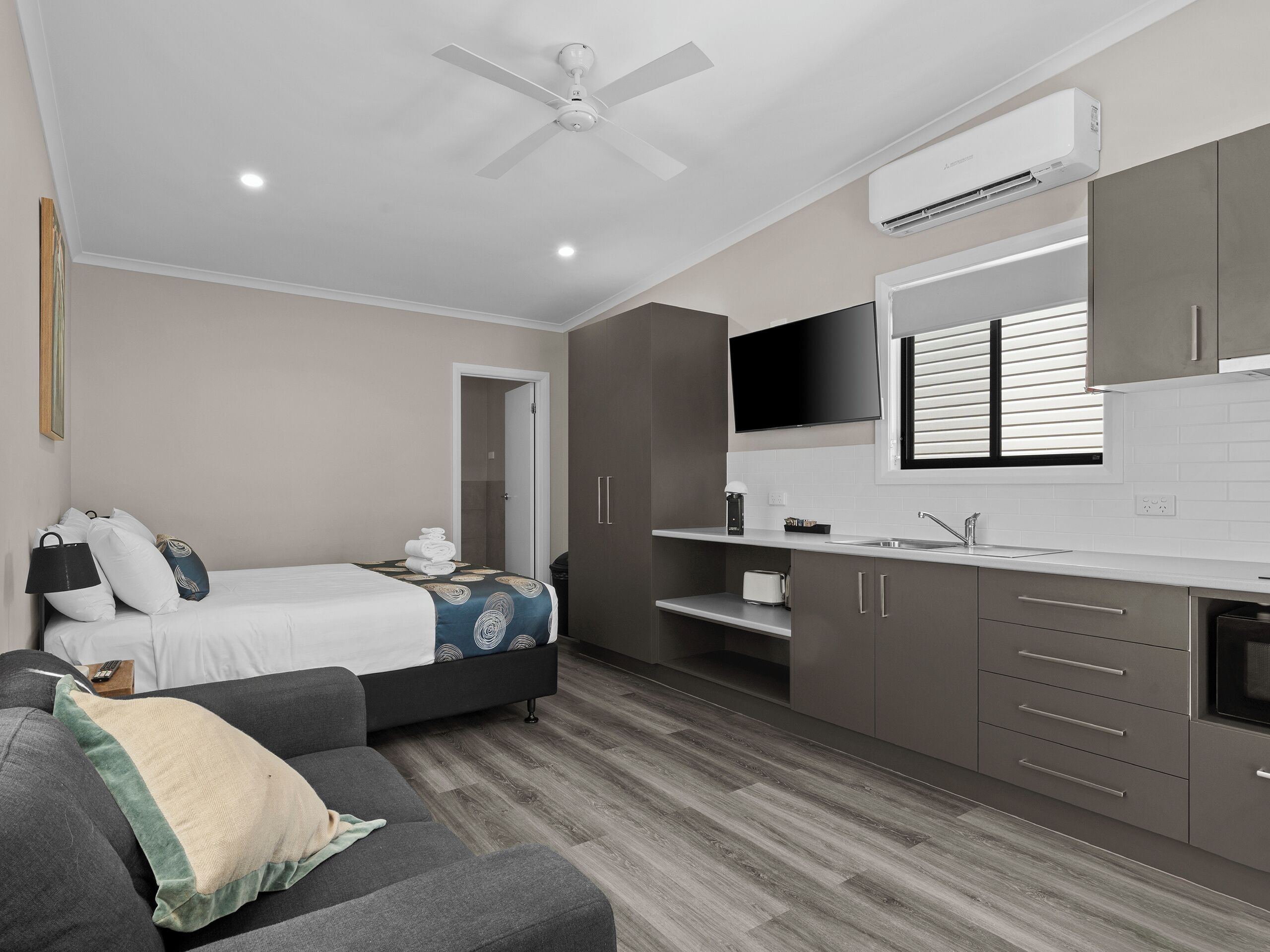 A modern hotel room with a bed, a sofa, a ceiling fan, an air conditioning unit, a TV, and a kitchenette.