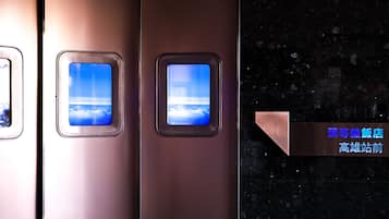 A metallic elevator interior with three windows displaying a blue sky and clouds. A sign indicates a restaurant and a high-rise building.