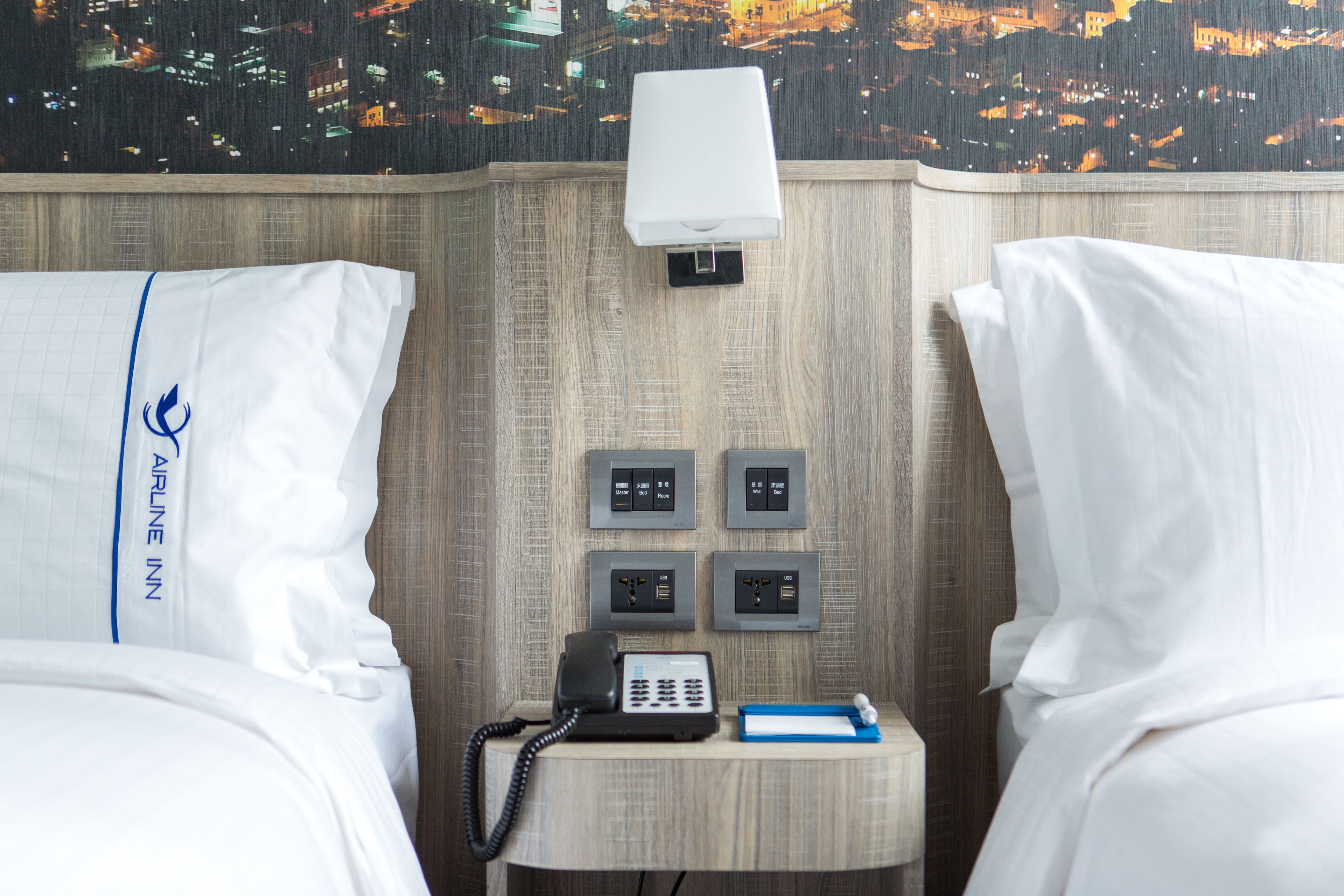 Habitación de hotel con dos camas, un teléfono en la mesita de noche, una pequeña mesa con un bloc de notas y bolígrafo, y un teléfono en la pared.