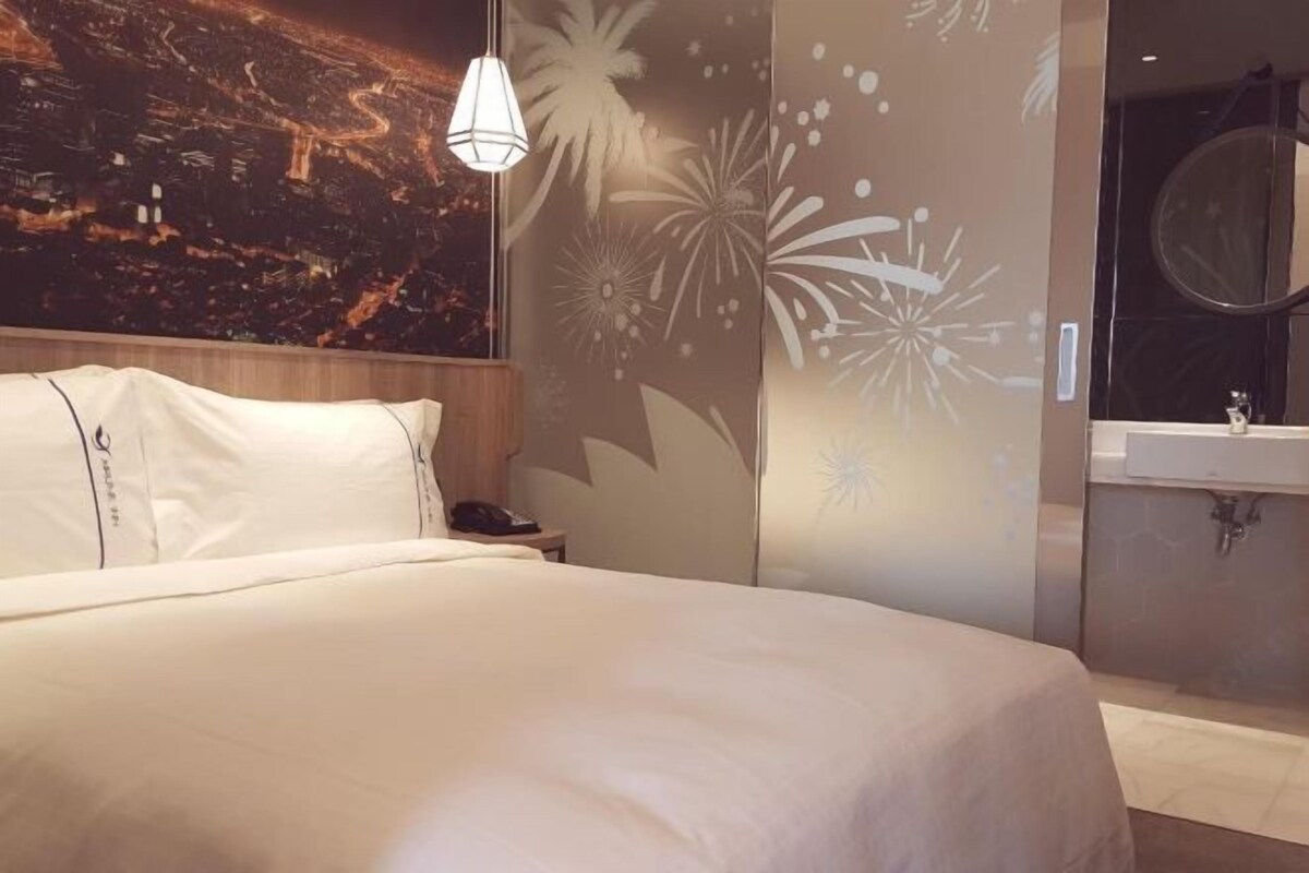 Quarto de hotel com cama, mesa de cabeceira, telefone fixado na parede, decoração floral e banheiro com pia e espelho.