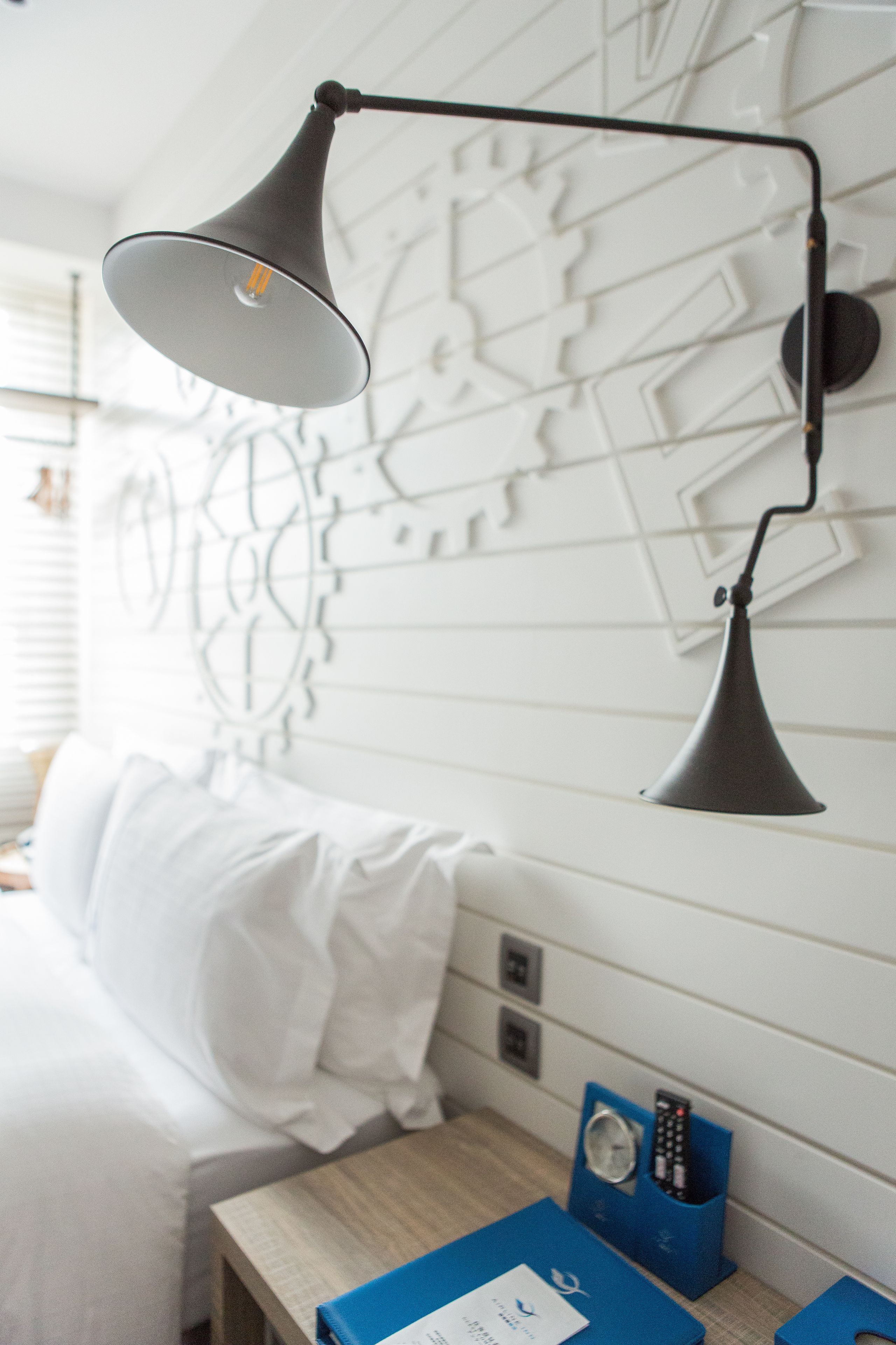 Een hotelkamer met een bed, een nachtkastje met een lamp, een muur met een geometrisch patroon en een stopcontact.