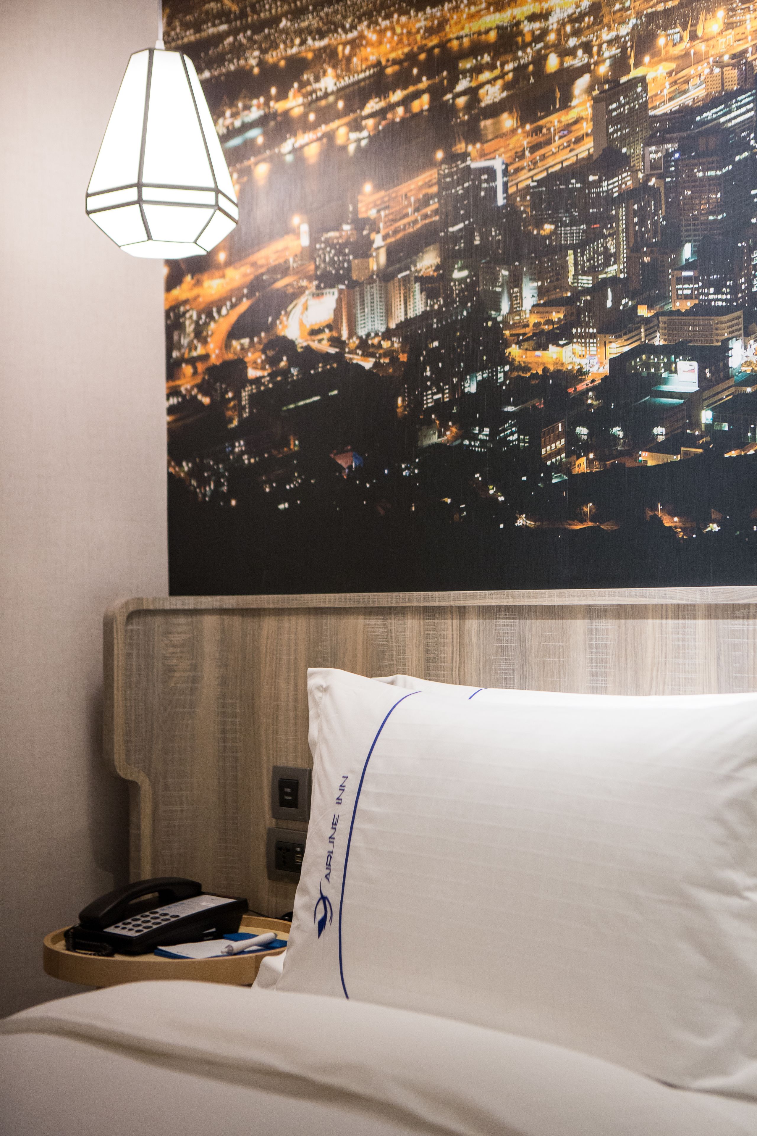 Habitación de hotel con vistas a la ciudad, cama con ropa blanca, mesita de noche con un teléfono y una libreta, y una lámpara colgante.