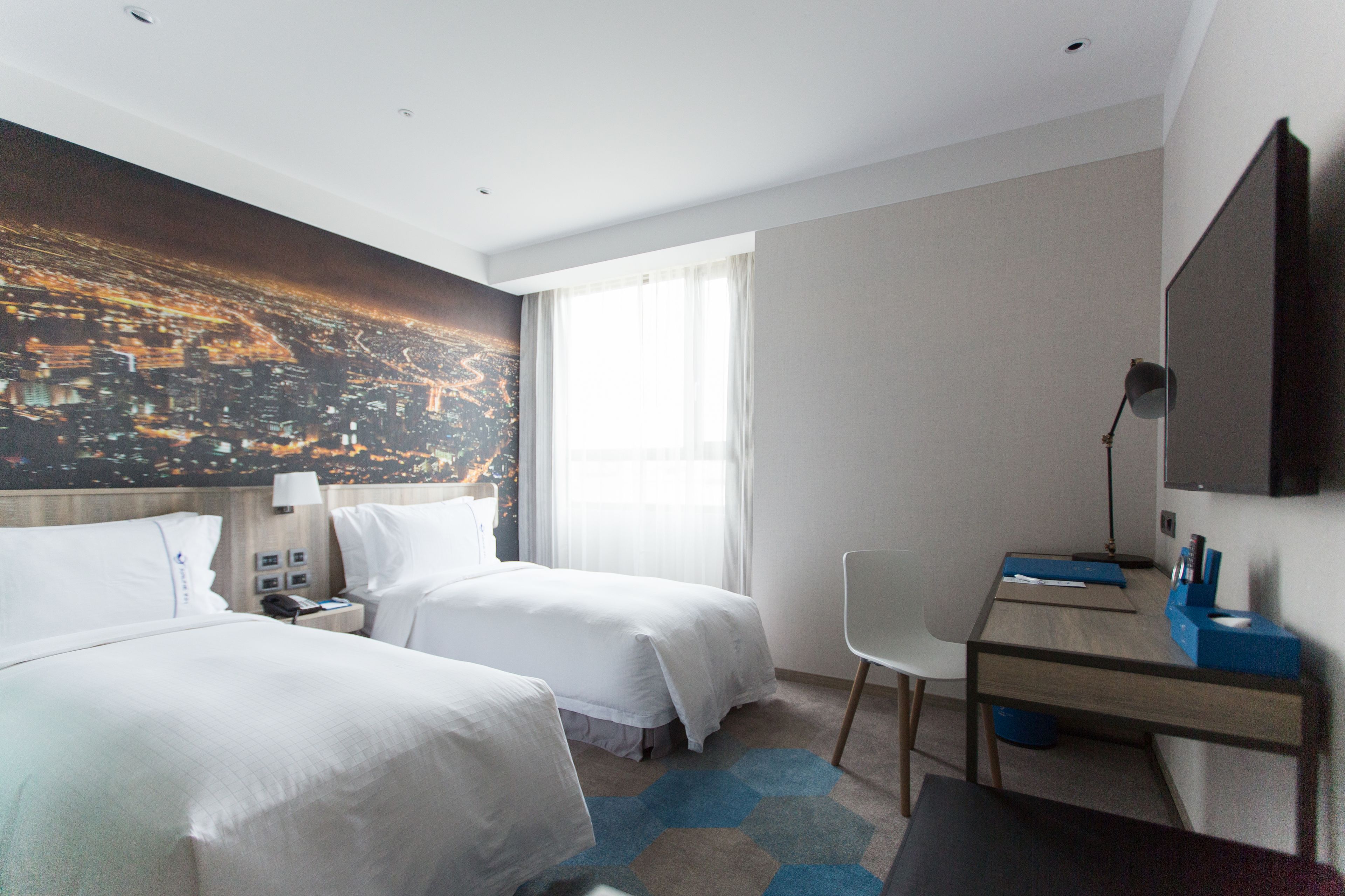 A hotel room with two beds, a desk, a chair, a TV, and a cityscape mural on the wall.