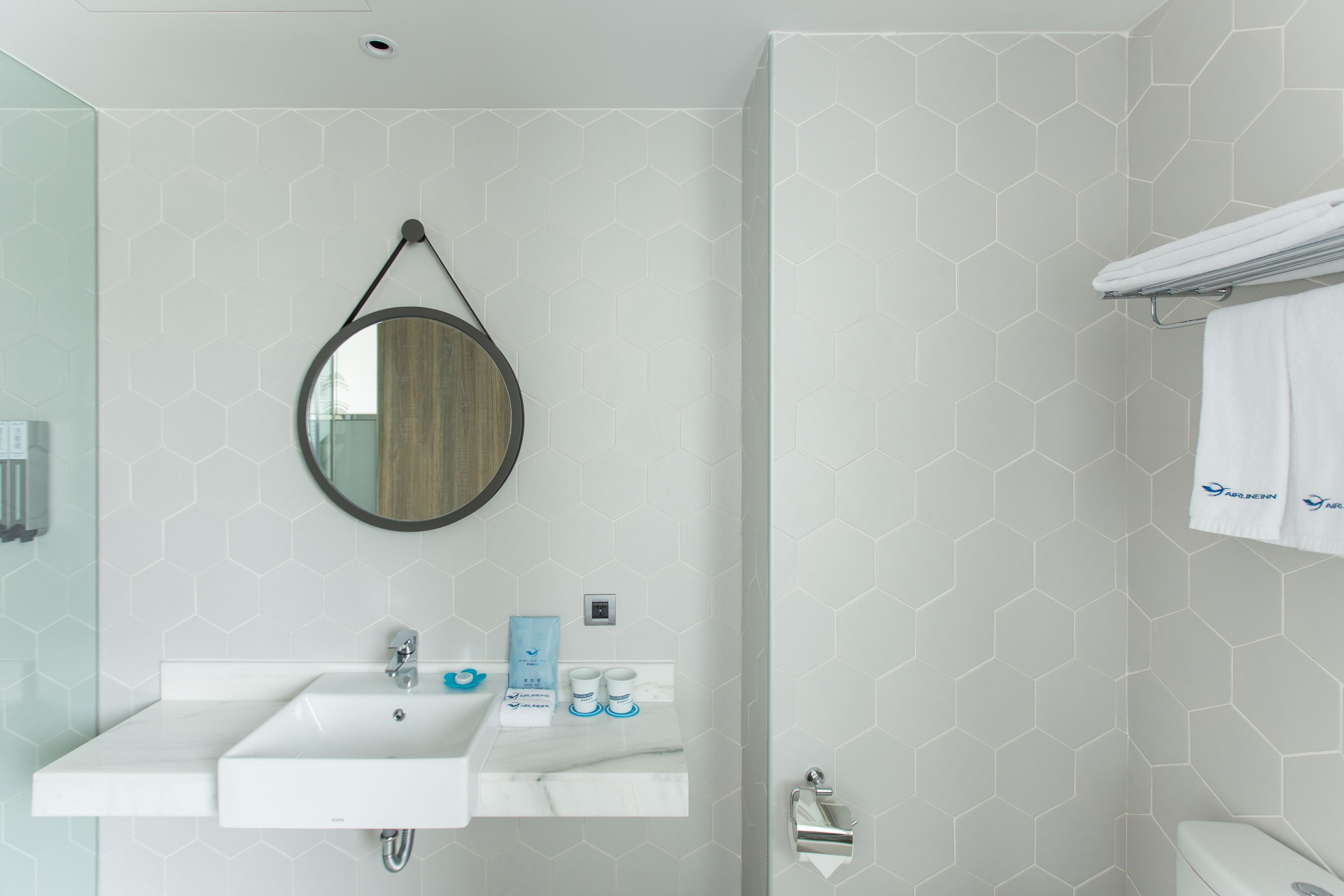 Un baño moderno con un lavabo blanco, espejo y inodoro. Hay artículos de aseo en el lavabo y un perchero sobre el inodoro.