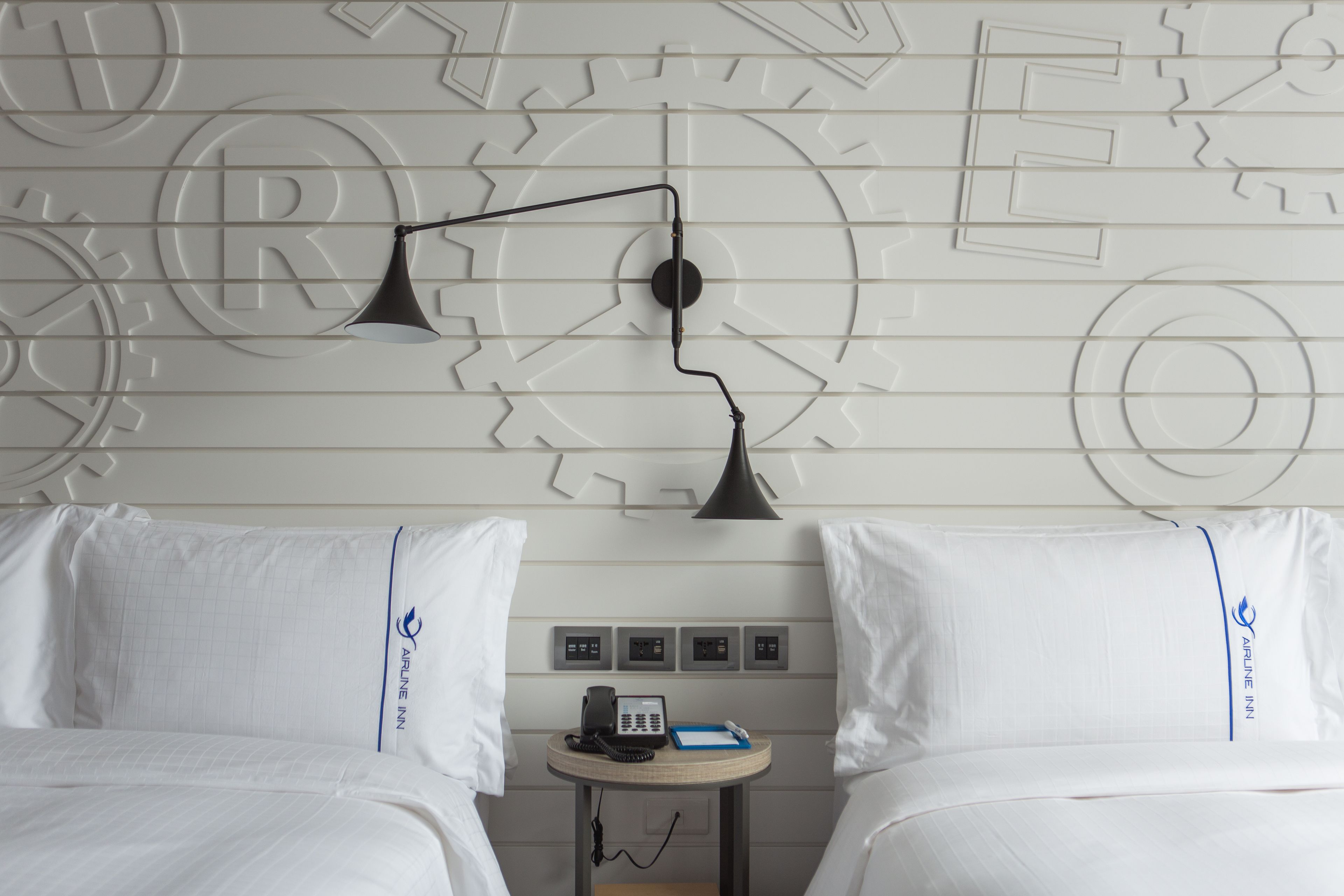 Une chambre d’hôtel avec deux lits, une petite table avec un téléphone et un bloc-notes, et un mur orné d’un motif représentant des engrenages.