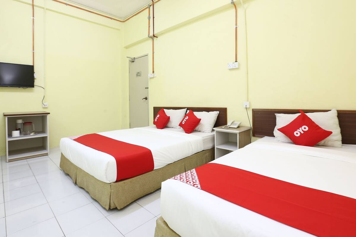 Habitación de hotel con dos camas, cada una con cubrecama y almohadas rojas y blancas. Hay una mesita de noche con un teléfono y un estante con artículos de tocador. Las paredes son de color amarillo claro y el techo es blanco.