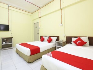 Quarto de hotel com duas camas, cada uma com colcha e almofadas vermelhas e brancas. Possui uma pequena mesa de cabeceira com um telefone e uma prateleira com produtos de higiene pessoal. O quarto tem paredes amarelo claro e teto branco.