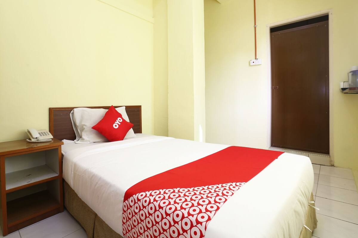Habitación de hotel con una cama, una mesita de noche con teléfono, un almohadón rojo y una colcha de cuadros rojos y blancos.
