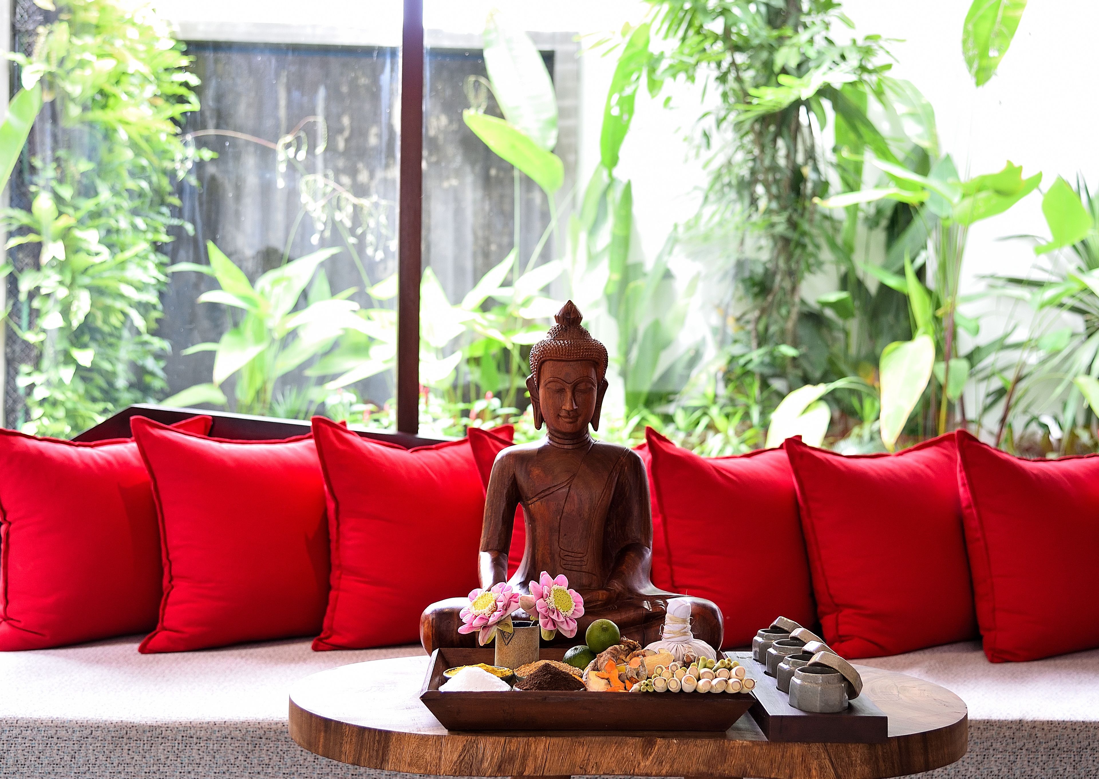 A Buddha statue on a wooden tray with offerings, surrounded by red cushions on a round table, with lush greenery in the background.