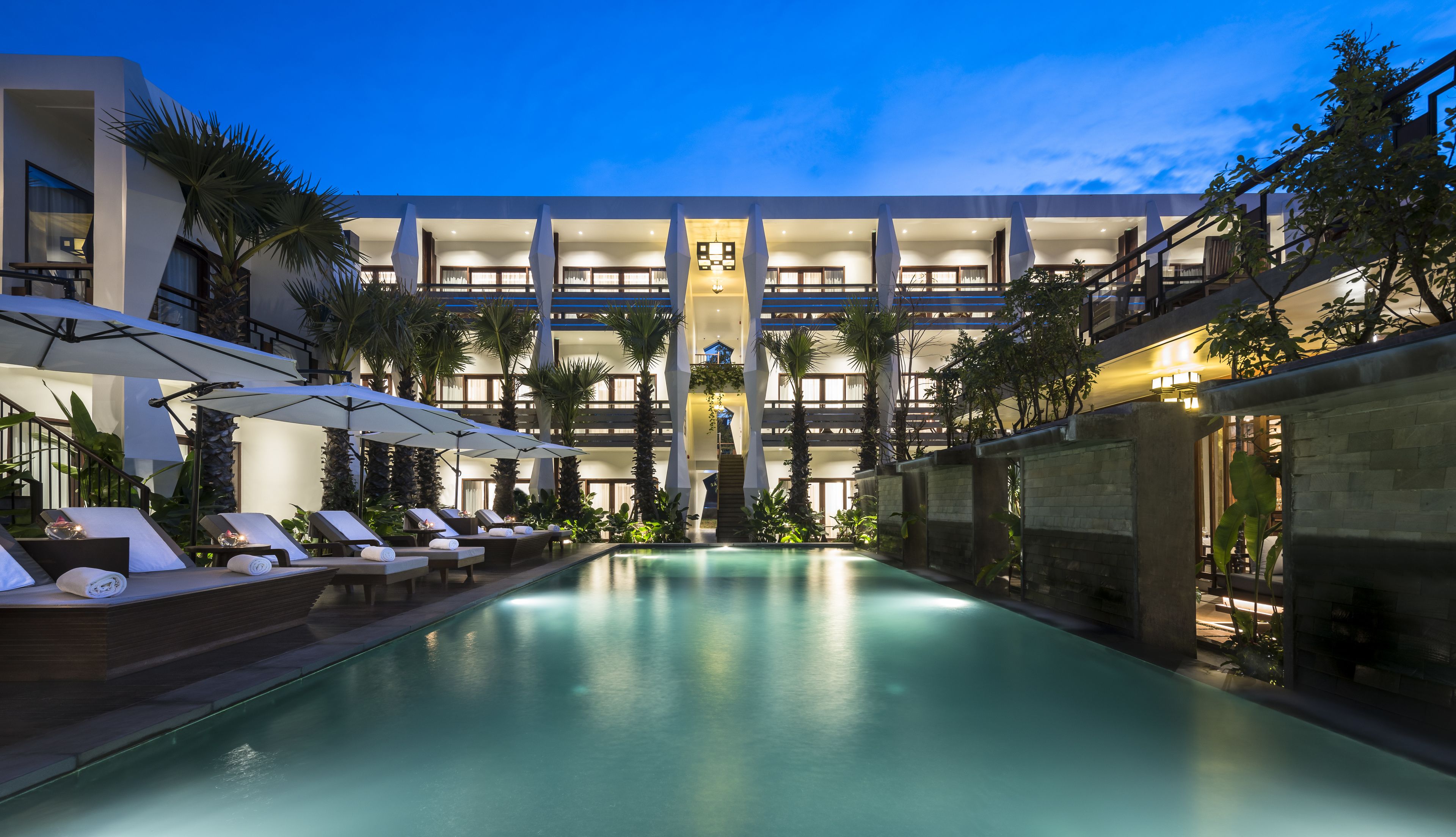 Un hotel moderno con piscina, varios pisos y zona de estar exterior con sombrillas.