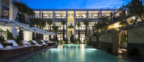 Un hotel moderno con piscina, varios pisos y zona de estar exterior con sombrillas.