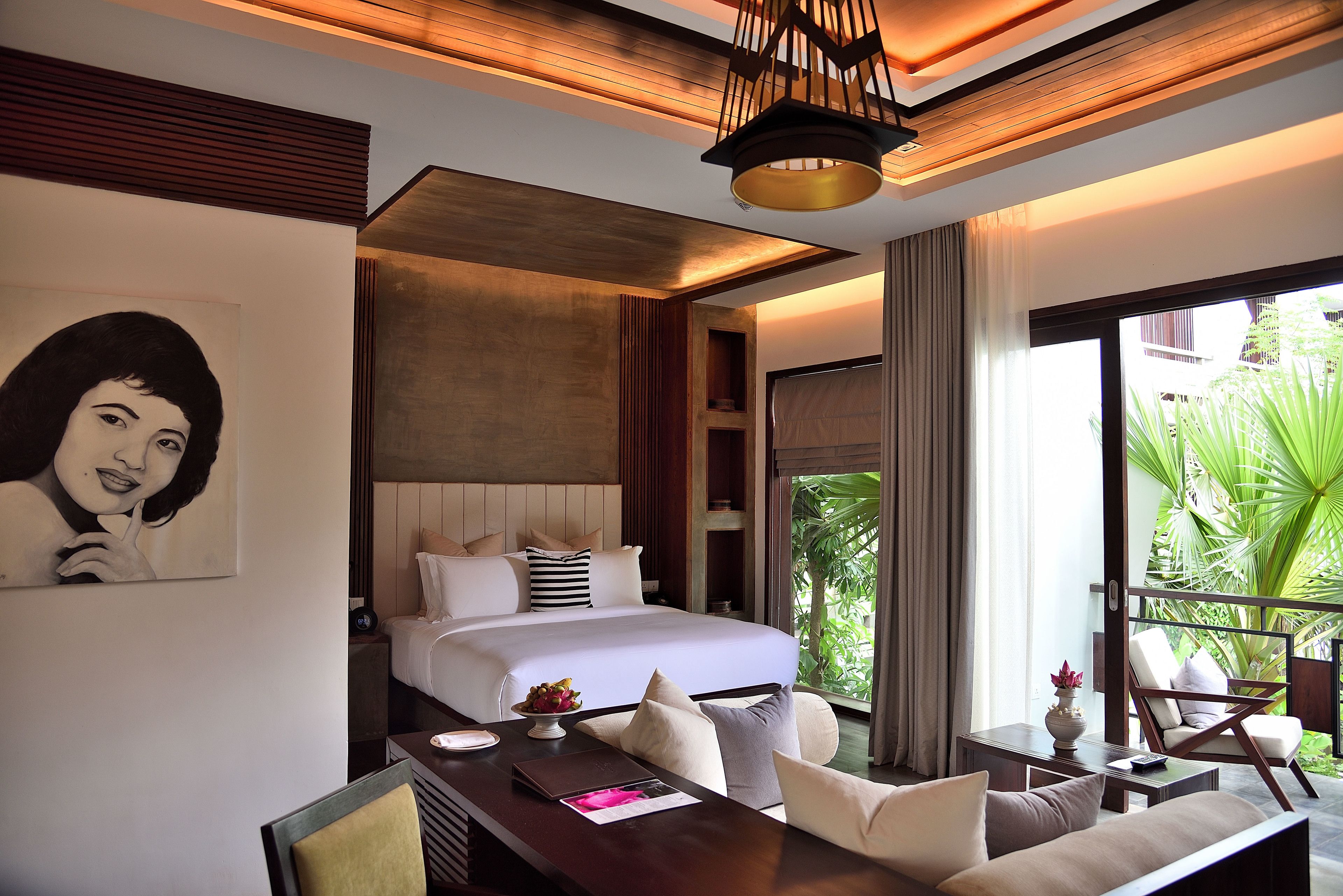 A modern hotel room with a large bed, a dining area, and a view of greenery outside.