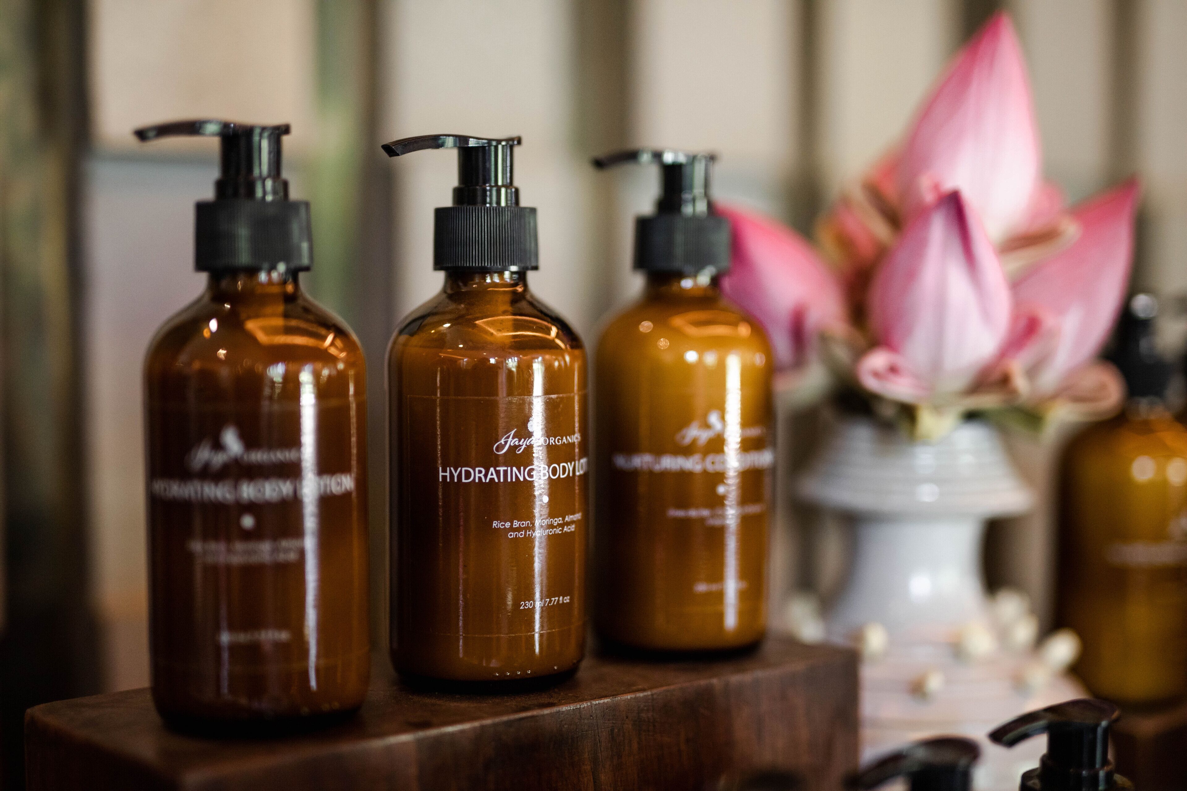 Tres botellas color ámbar con dosificadores negros, etiquetadas como “HYDRATING BODY LOTION”, sobre una superficie de madera con una flor de loto rosa de fondo.
