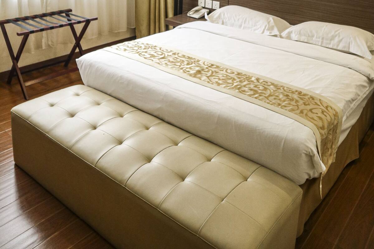 A hotel room with a large bed, a bedside table with a lamp, a wooden bench, and a window with curtains.