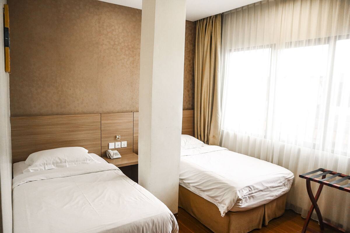 A hotel room with two beds, a wooden headboard, a small table with a phone, and a window with sheer curtains.