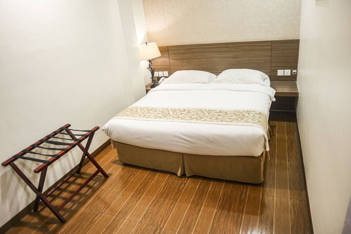 A hotel room with a wooden floor, a bed with white bedding, a nightstand with a lamp, and a wooden drying rack.