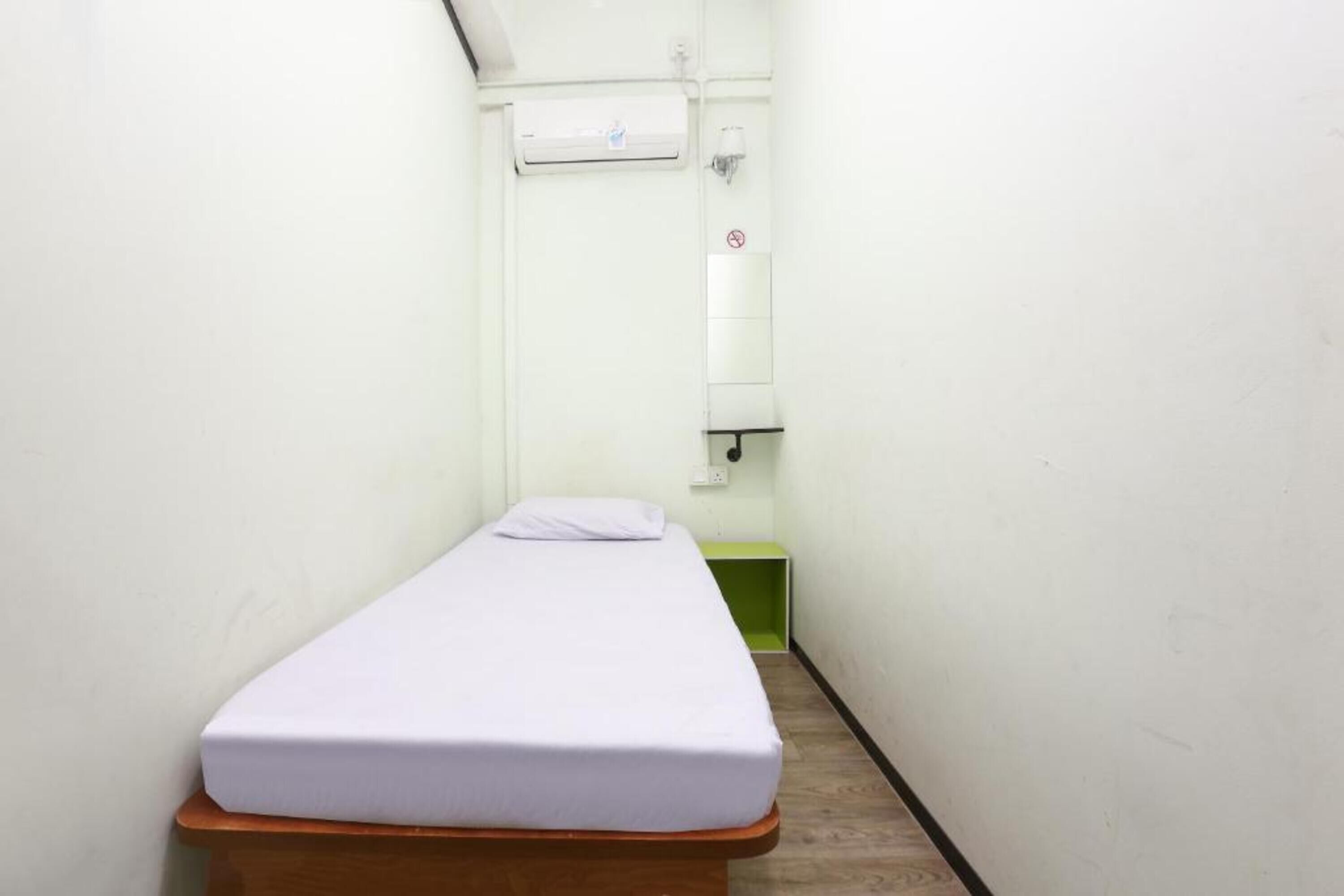 A narrow room with a single bed, a green storage unit, and a wall-mounted air conditioning unit.