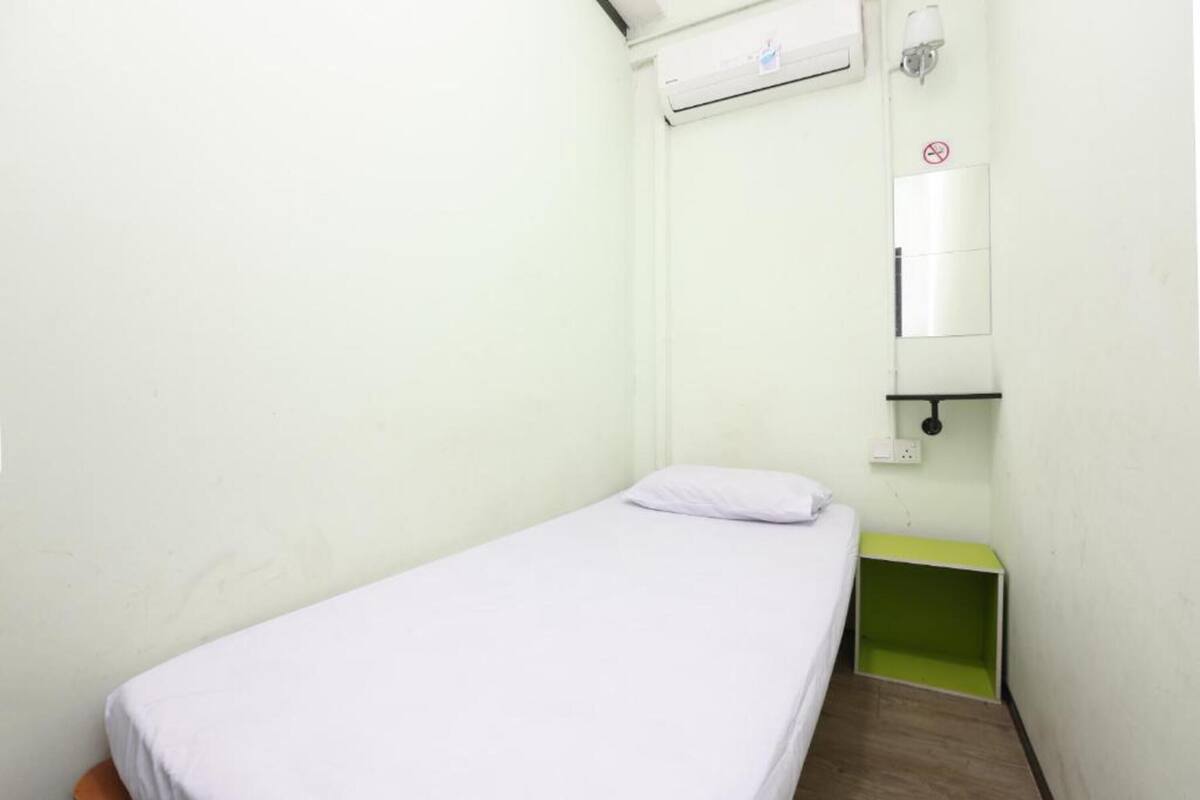A small, single-bed room with a white bedspread, a green nightstand, a wall-mounted air conditioner, and a mirror with a no-smoking sign.
