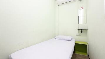 A small, single-bed room with a white bedspread, a green nightstand, a wall-mounted air conditioner, and a mirror with a no-smoking sign.