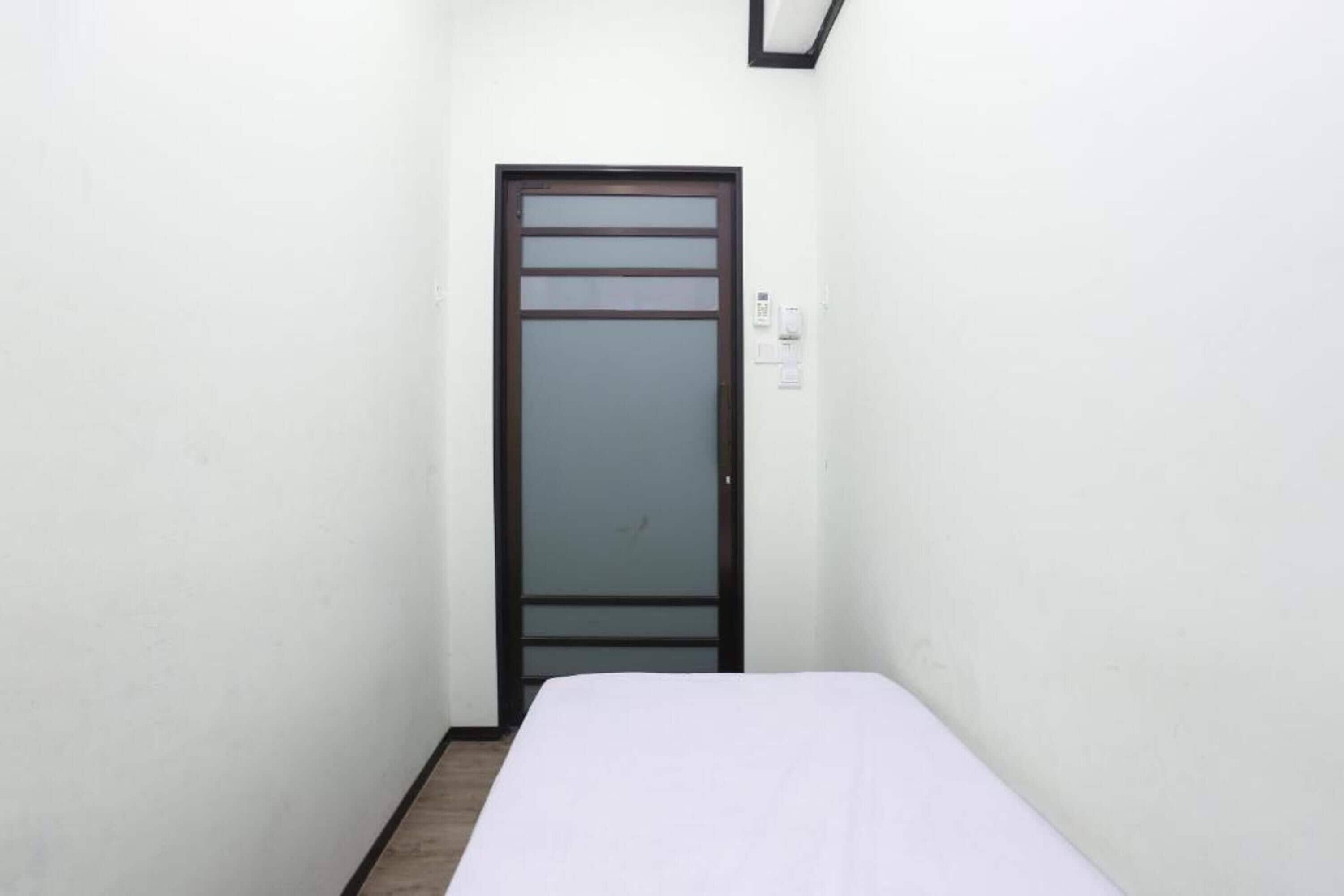Um quarto pequeno e estreito, com uma cama de solteiro, uma porta com painel de vidro fosco e um interruptor de luz na parede.