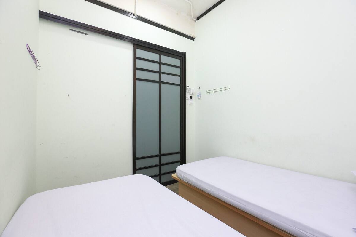 A small, clean room with two single beds, a sliding door, and a simple wall-mounted light fixture.