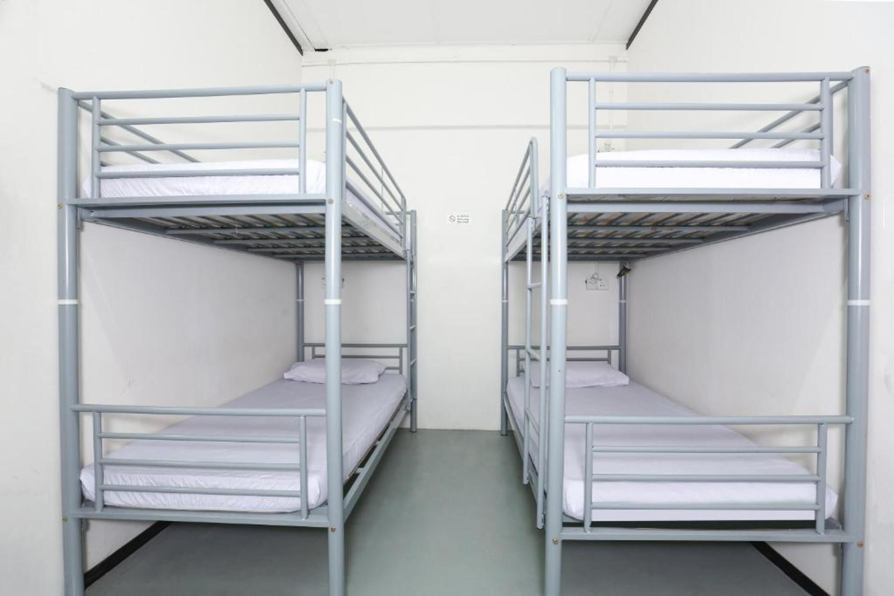 Two bunk beds in a narrow room with a plain white wall and a visible electrical outlet.